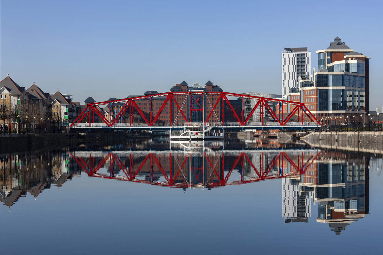 Calm waters reflecting the vibrant red bridge at Salford Quays, with modern buildings and clear blue skies, showcasing Manchester's mix of industrial heritage and contemporary architecture